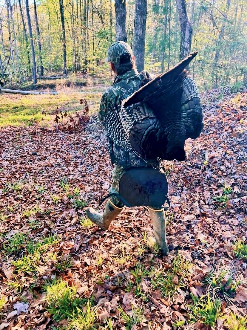 Hunter carries a large, harvested turkey through a wooded area.