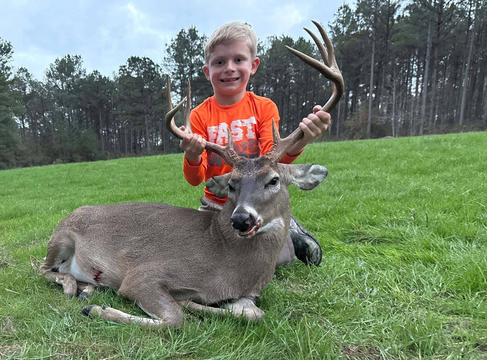 Young person holding antlers of a deer lying on green grass, smiling with an orange shirt outdoors.