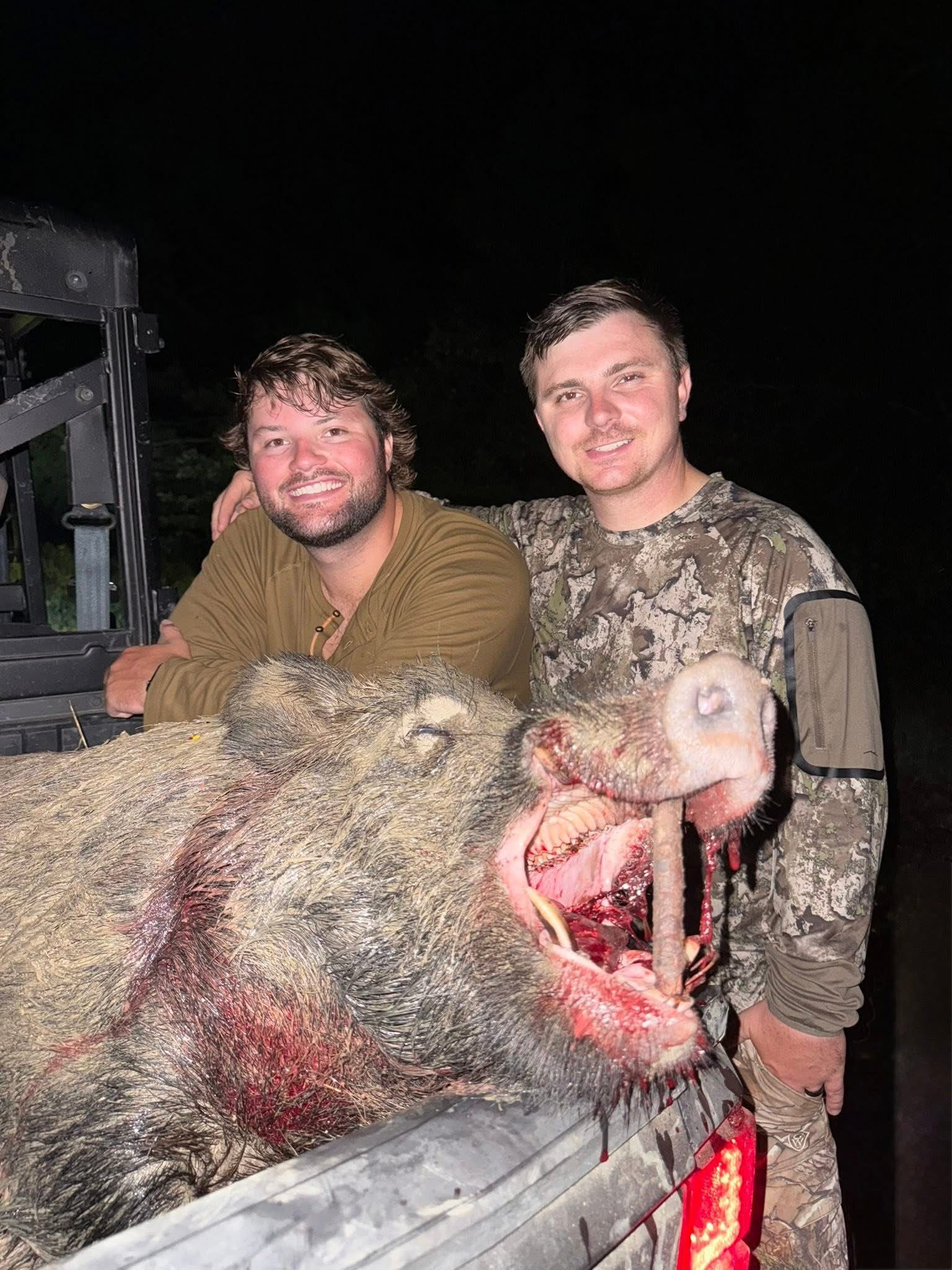 Two men pose with a large, dead wild hog at night. The hog has a bloody mouth.