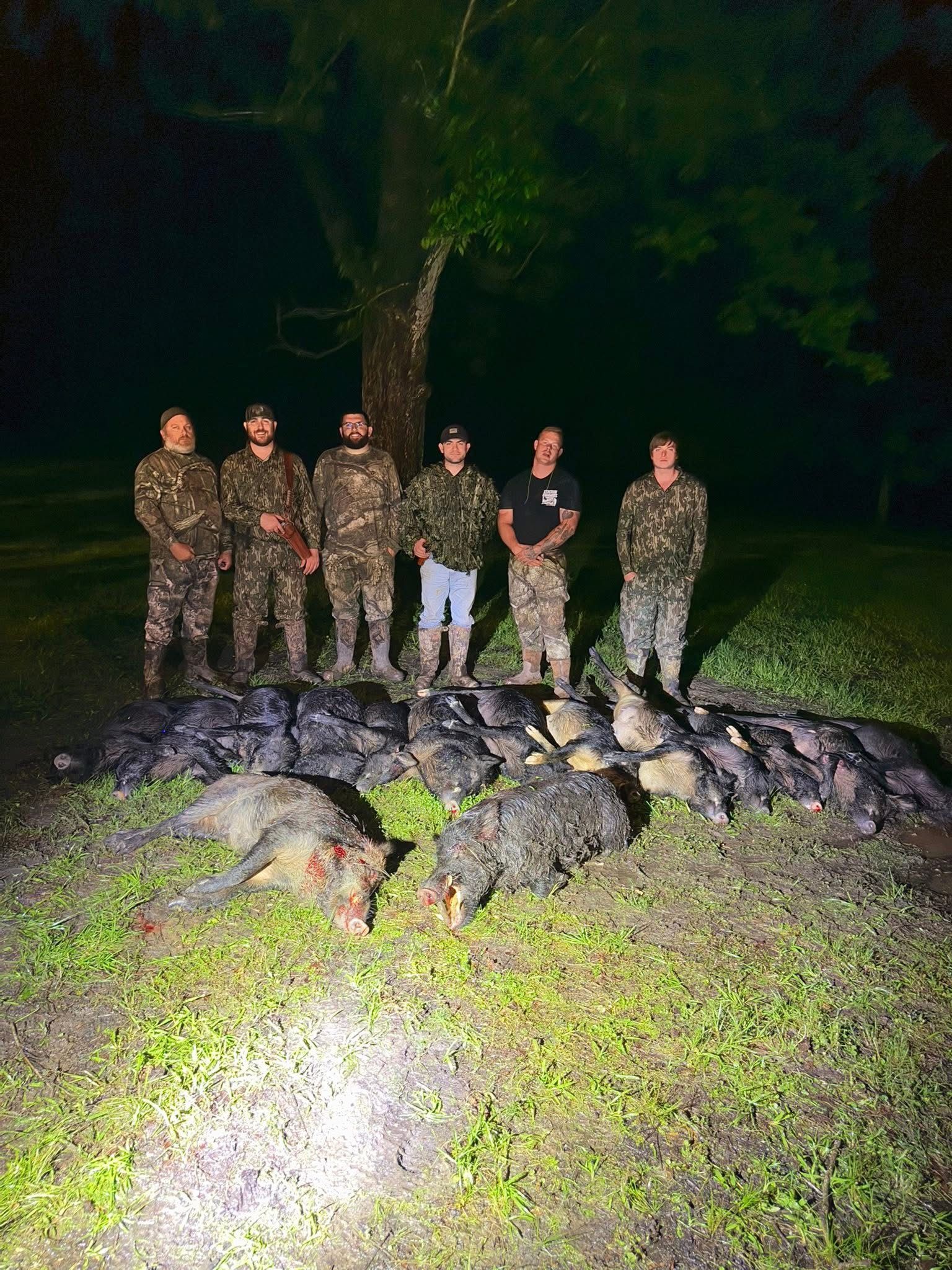 Six people in camo stand over several dead wild pigs at night.