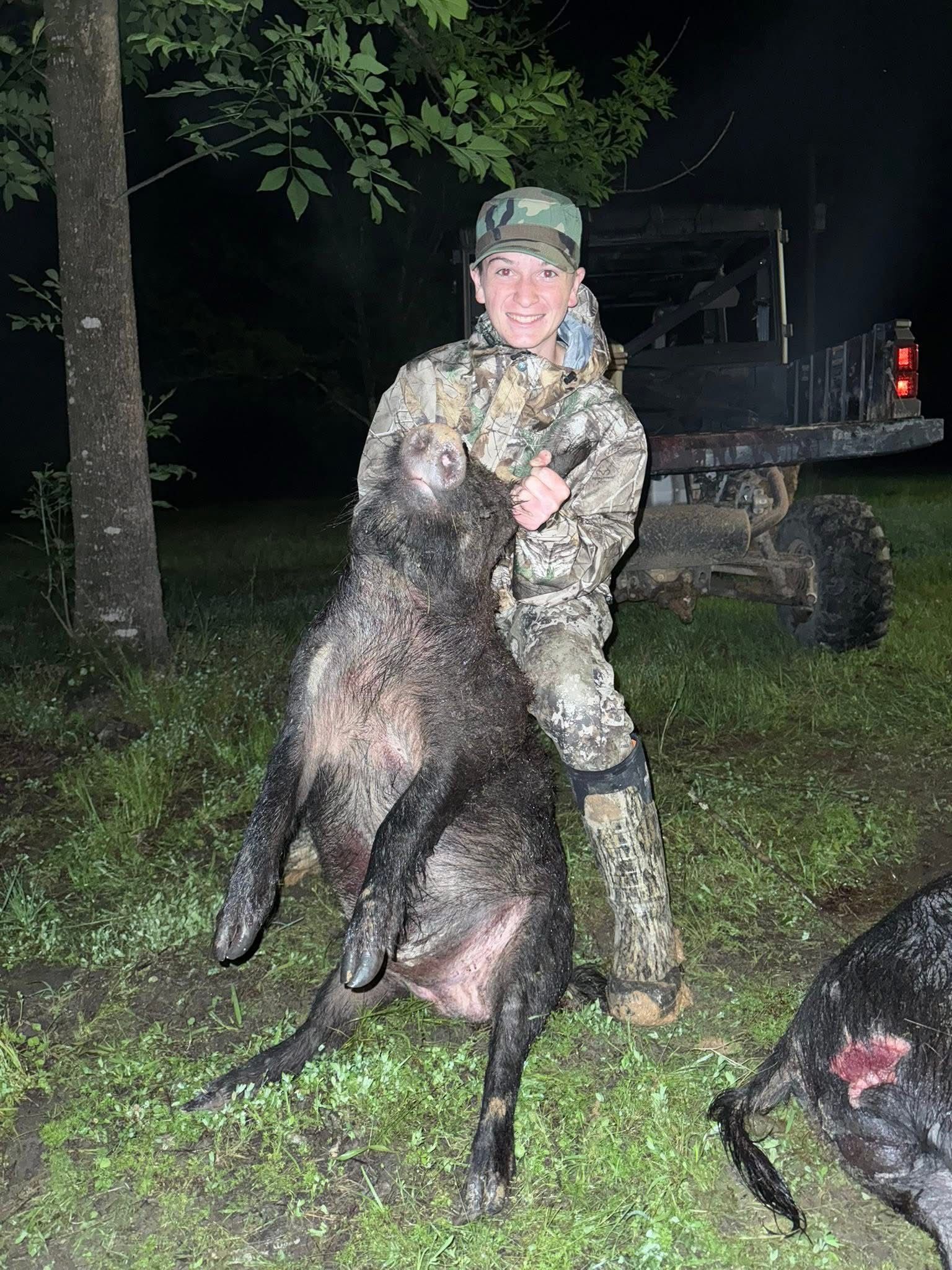 Young person in camo holding up a large, dark-colored wild pig in front of an ATV at night.
