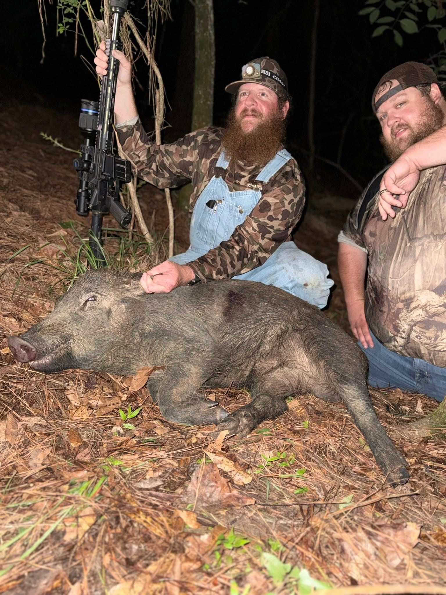 Two men pose with a dead wild boar in a wooded area. One holds a bow.