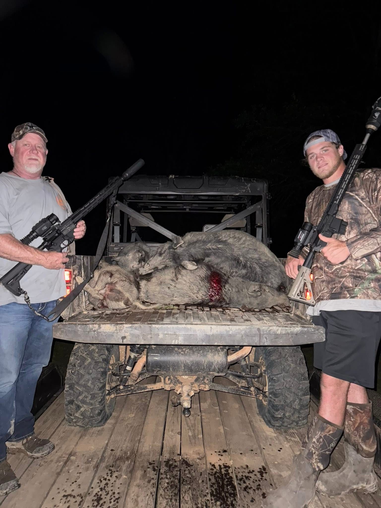Two men with rifles stand by a utility vehicle with several dead wild pigs. Dark setting.