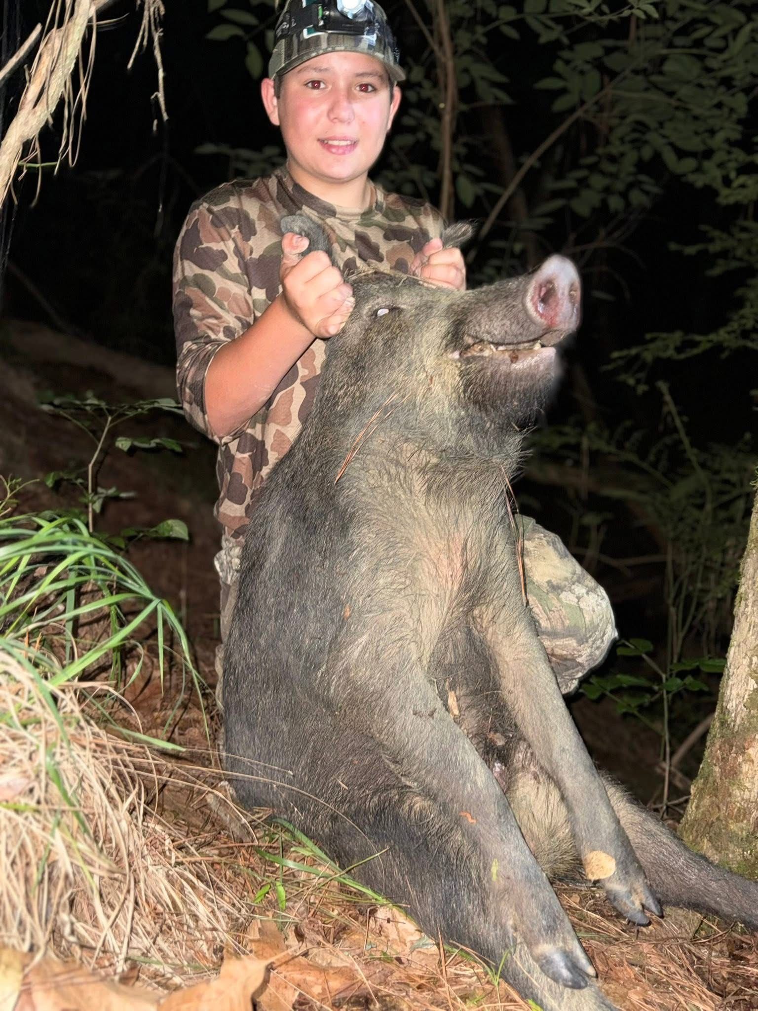 Boy in camouflage holds up a large, dead wild pig in a wooded area.