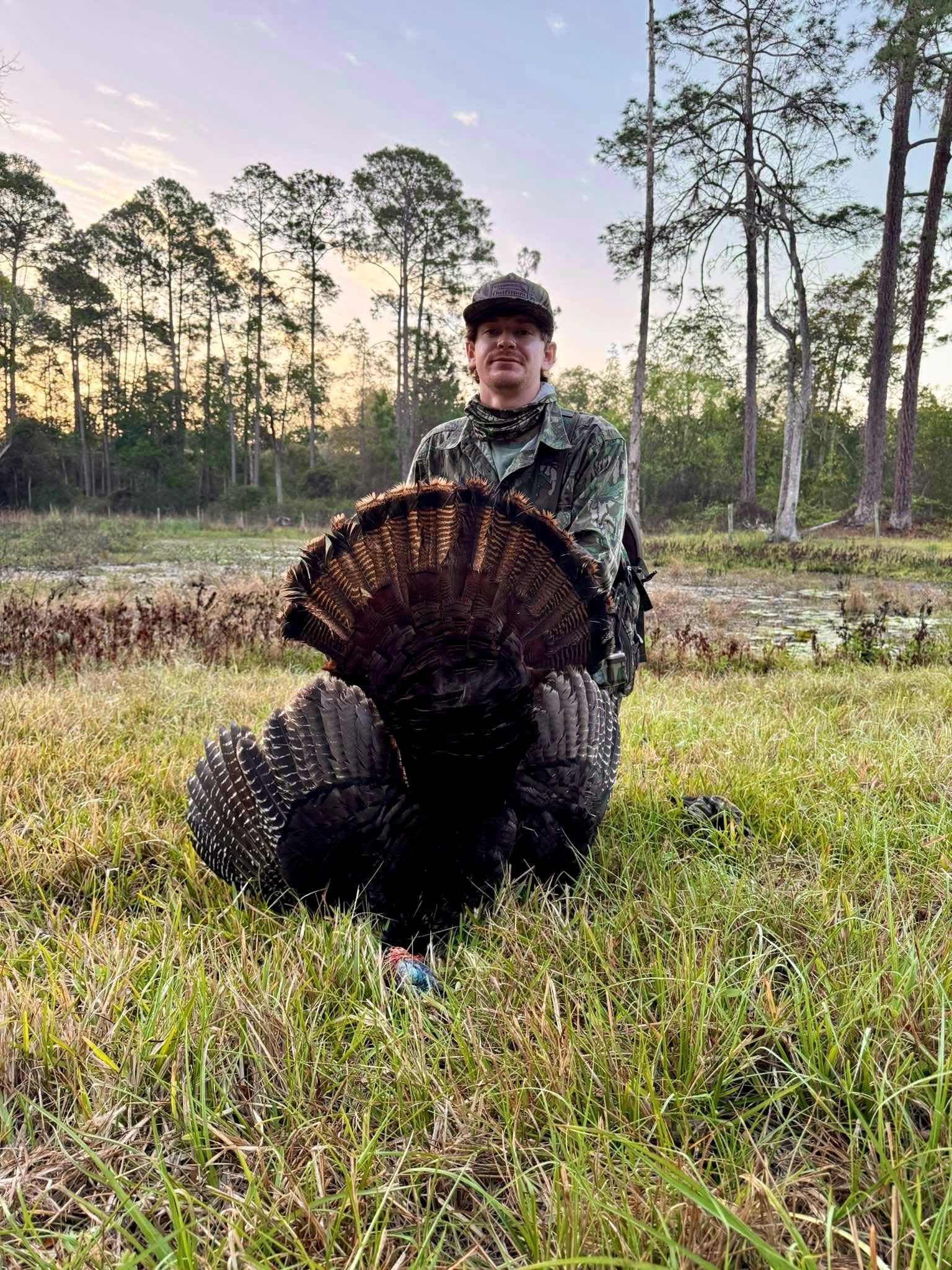 Hunter kneeling with a wild turkey in a field; trees in the background.