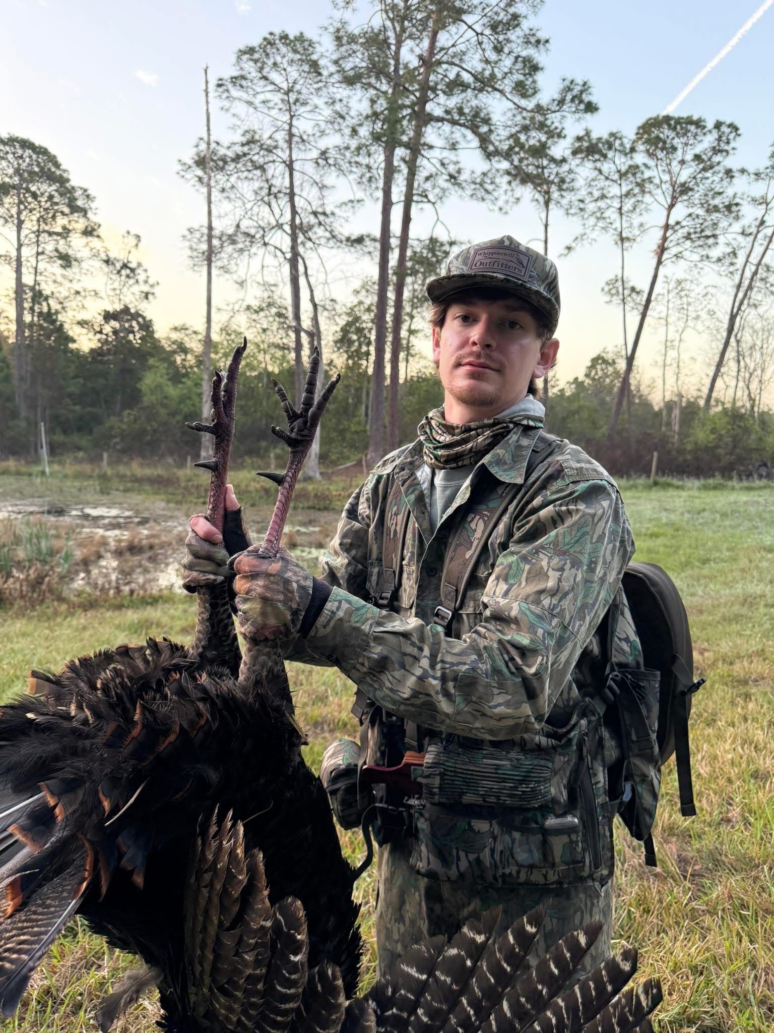 Man in camo holding a dead turkey in a field.