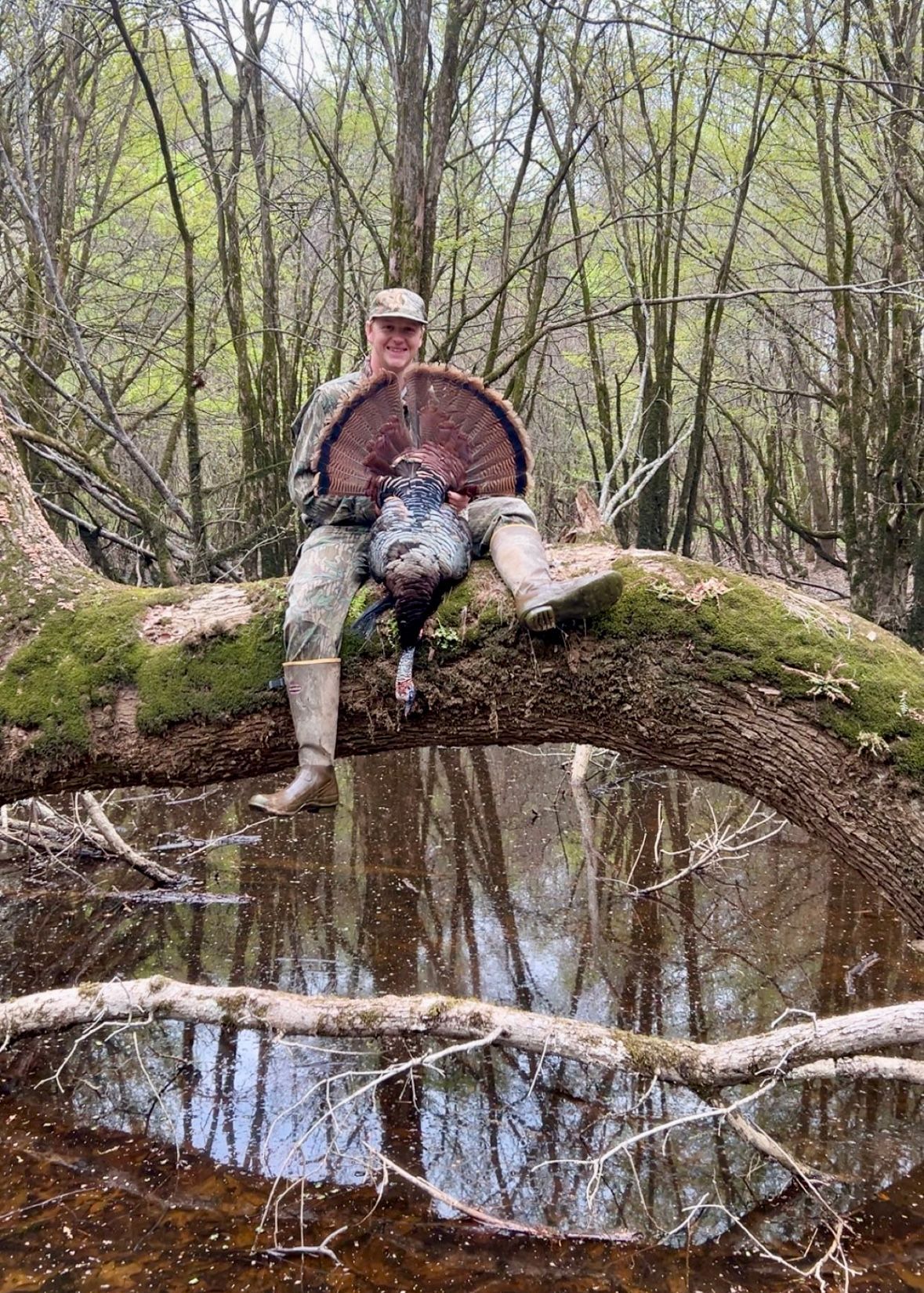 Hunter sits on log over water, holding a dead turkey, surrounded by trees.