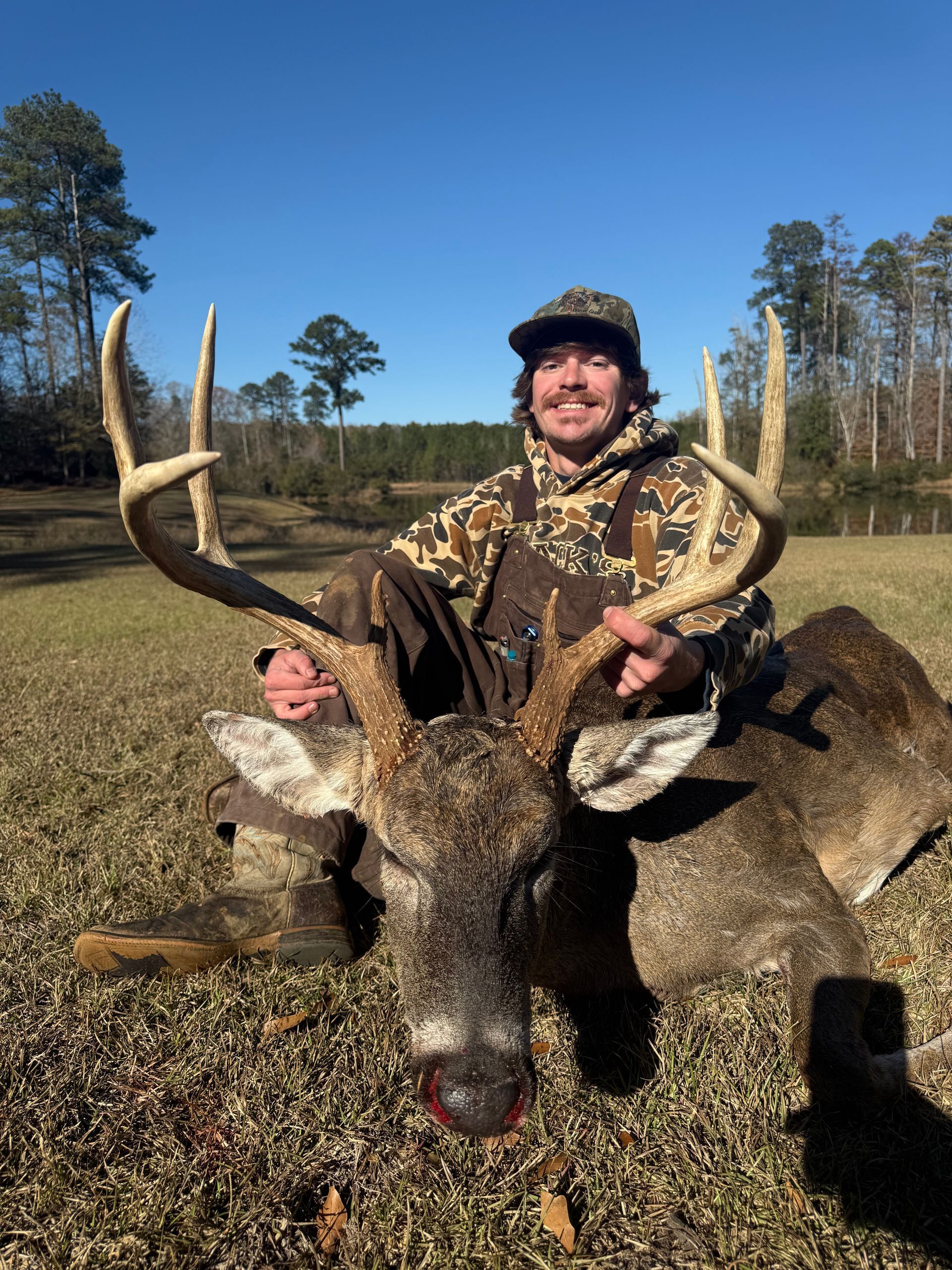 Man in camo, crouched, holds large deer antlers. Deer lies on ground, open field, bright blue sky.
