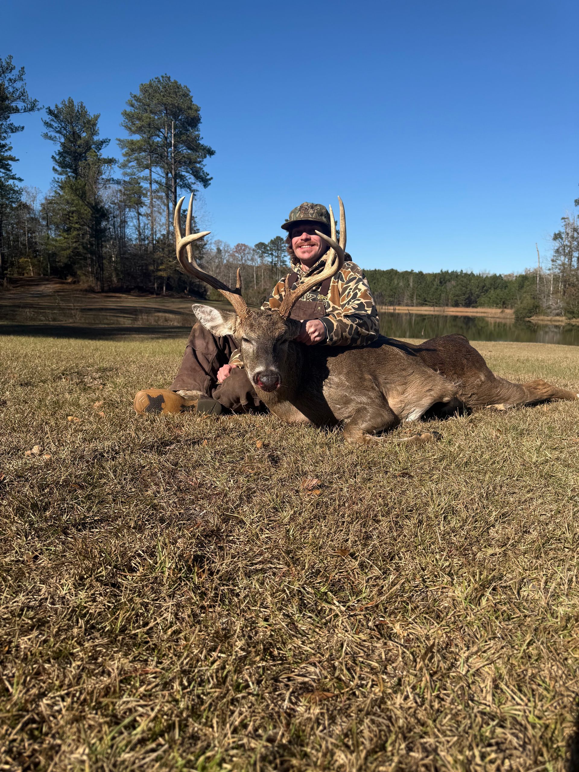 Man in camo with a large buck deer on a grassy field, under a blue sky.