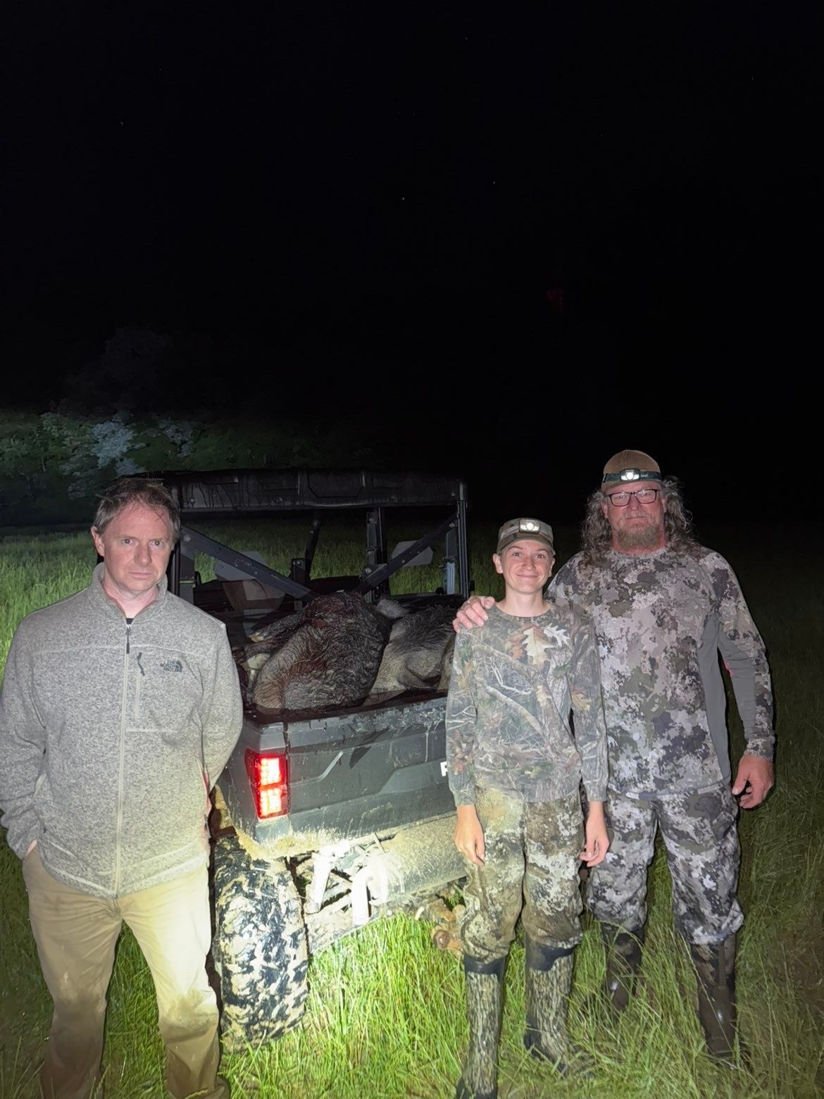 Three people stand by an ATV at night; the vehicle bed holds large game. The men wear camouflage; one holds a flashlight.