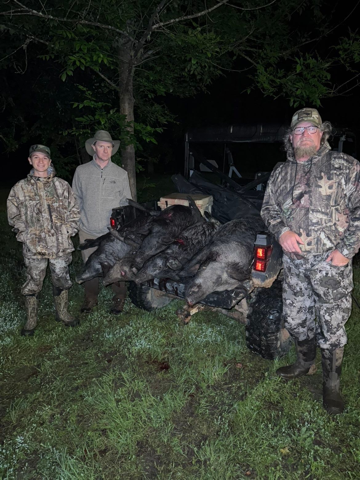 Three people pose with harvested animals near a utility vehicle at night.