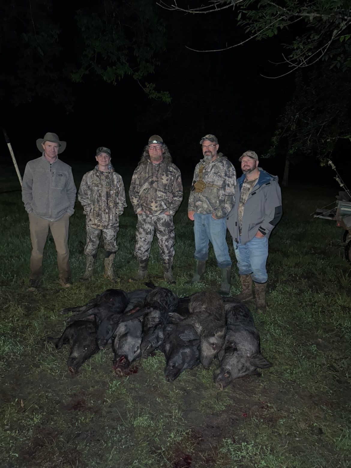 Five people with multiple wild pigs on the ground, likely after a hunt, in a grassy area at night.