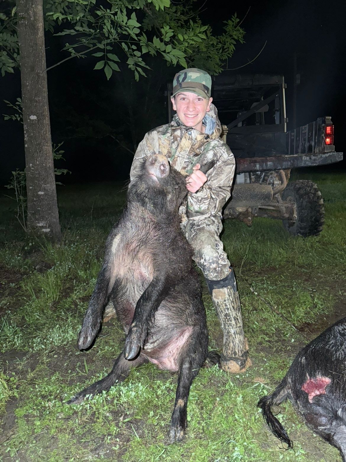 A person in camo holds up a large, dark-furred wild pig outdoors at night; a utility vehicle is in the background.