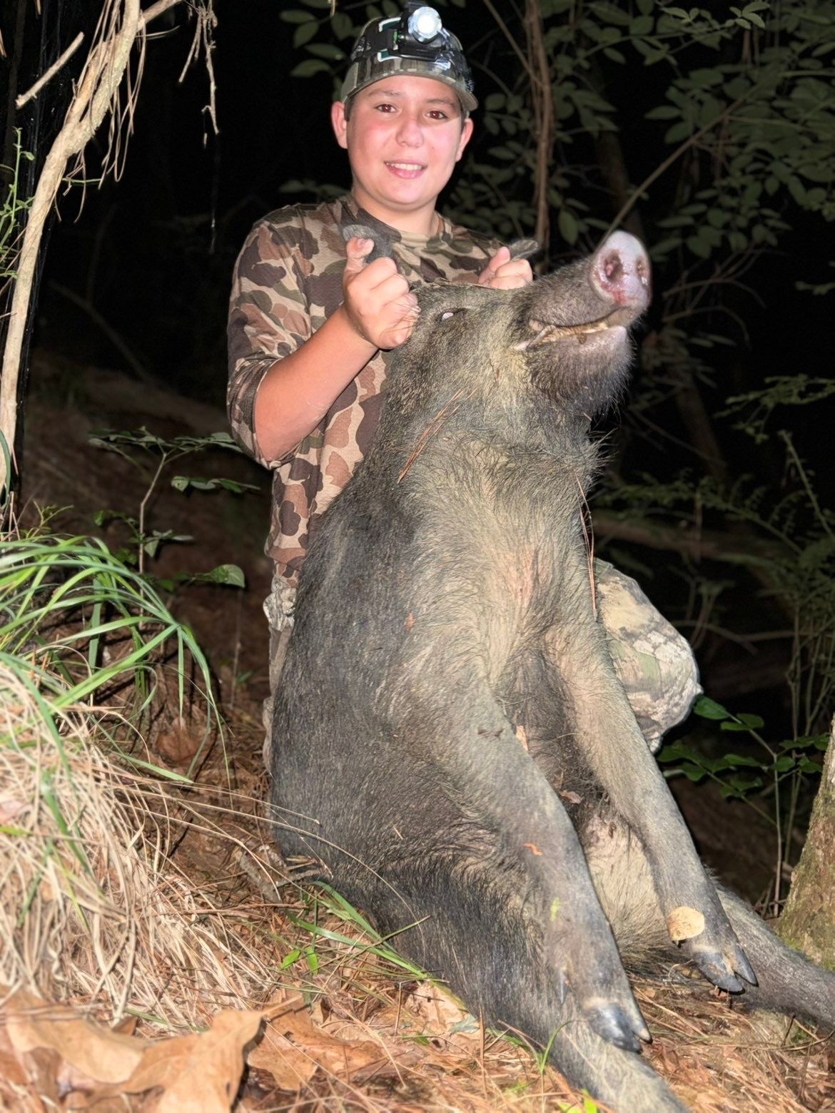 Boy in camouflage holds up a large, dead boar, thumbs up, in a dark outdoor setting.