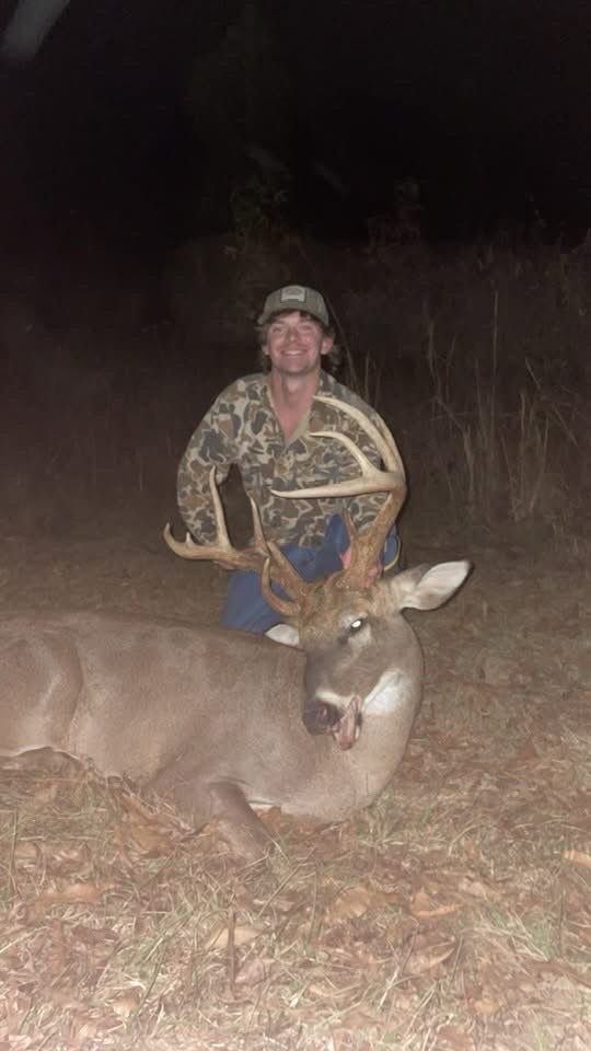Man kneeling beside a harvested buck at night, holding its antlers. He's wearing camo.