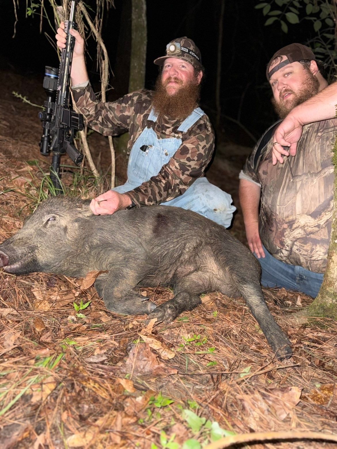 Two men pose with a dead wild hog in a forest. One man holds a bow; both are smiling.
