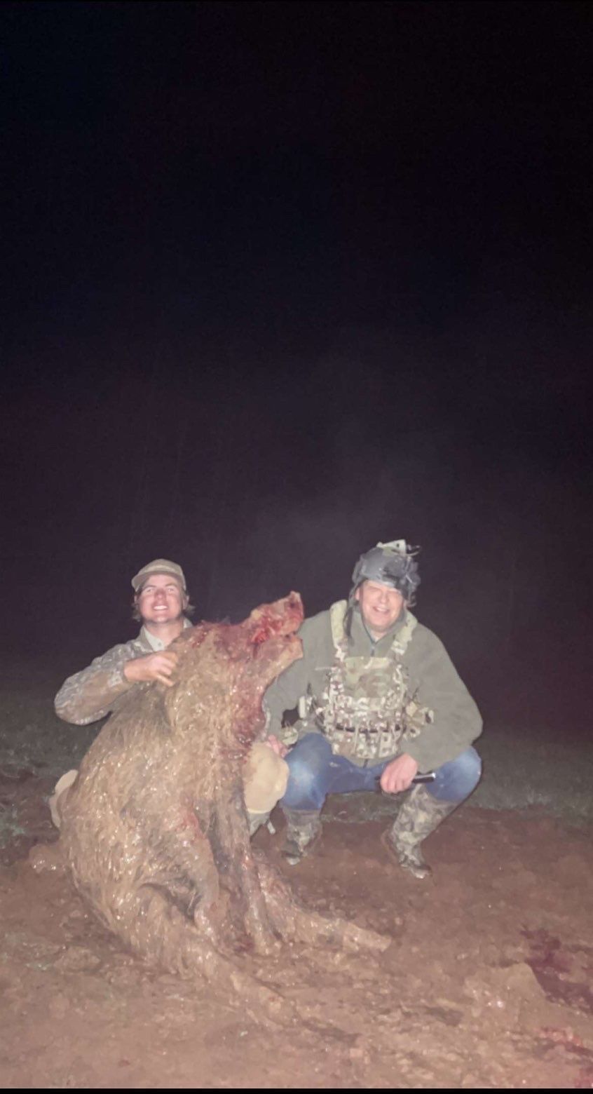 Two people kneel beside a dead boar at night. Both wear hats and hunting gear.