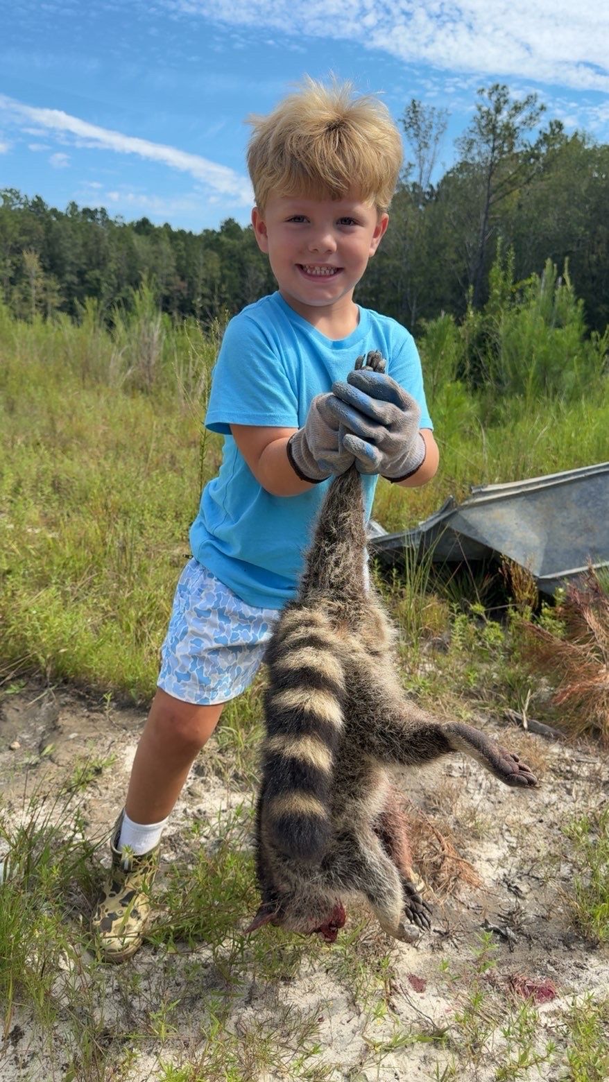 Young boy holding a dead, striped animal outdoors, wearing a blue shirt and shorts, smiling.
