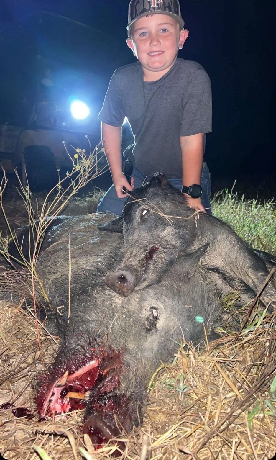 Boy kneels next to a dead hog at night. The animal's face shows blood, and the boy smiles.