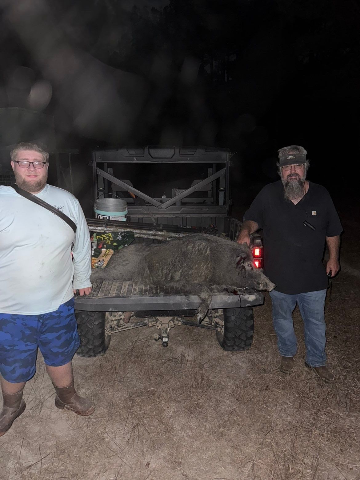 Two men stand by a utility vehicle at night. One wears a beard and hat. The other has glasses and shorts.