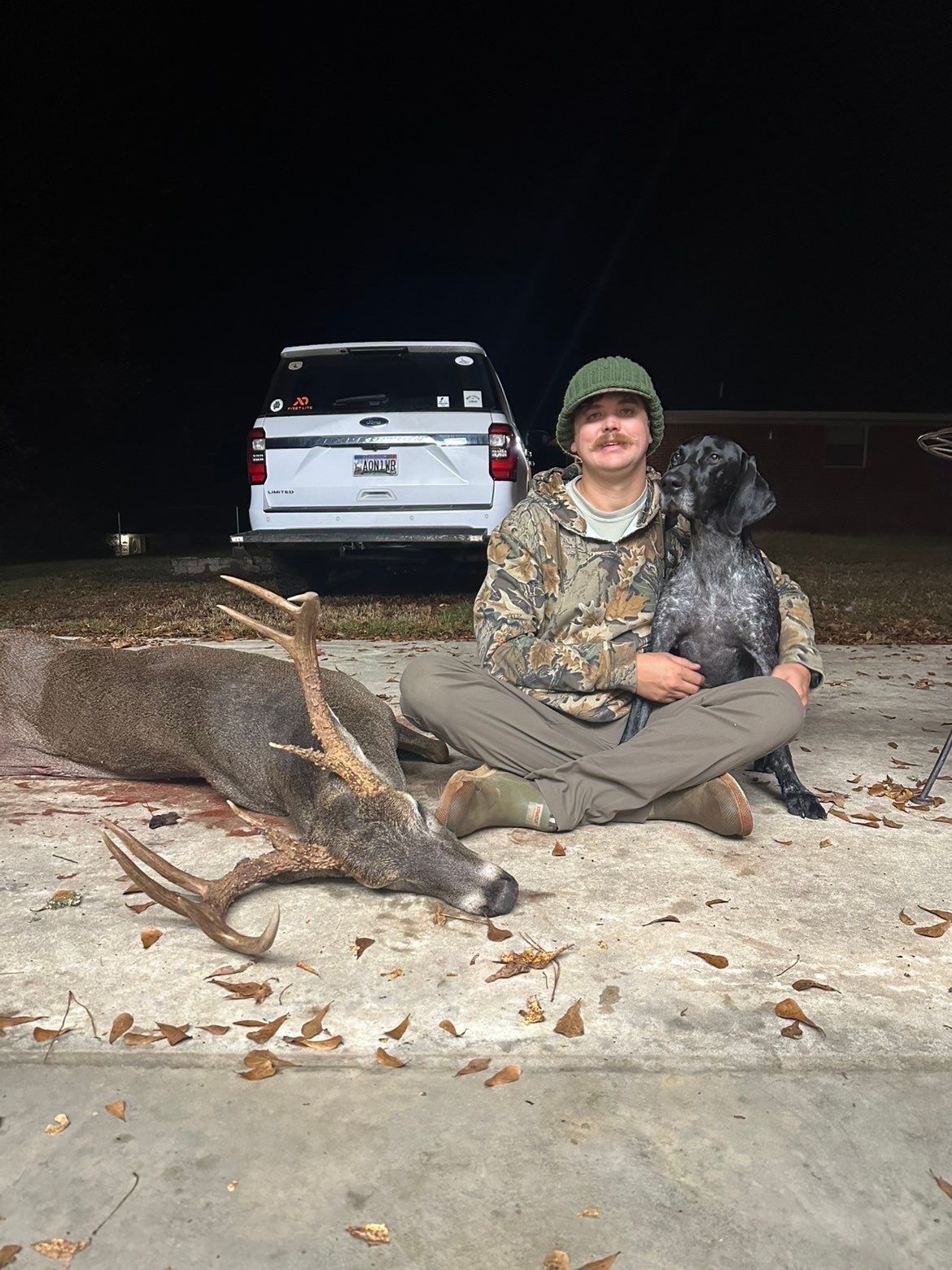 Man with dog sits beside a harvested deer. Dark setting; truck in background. The man wears camo.