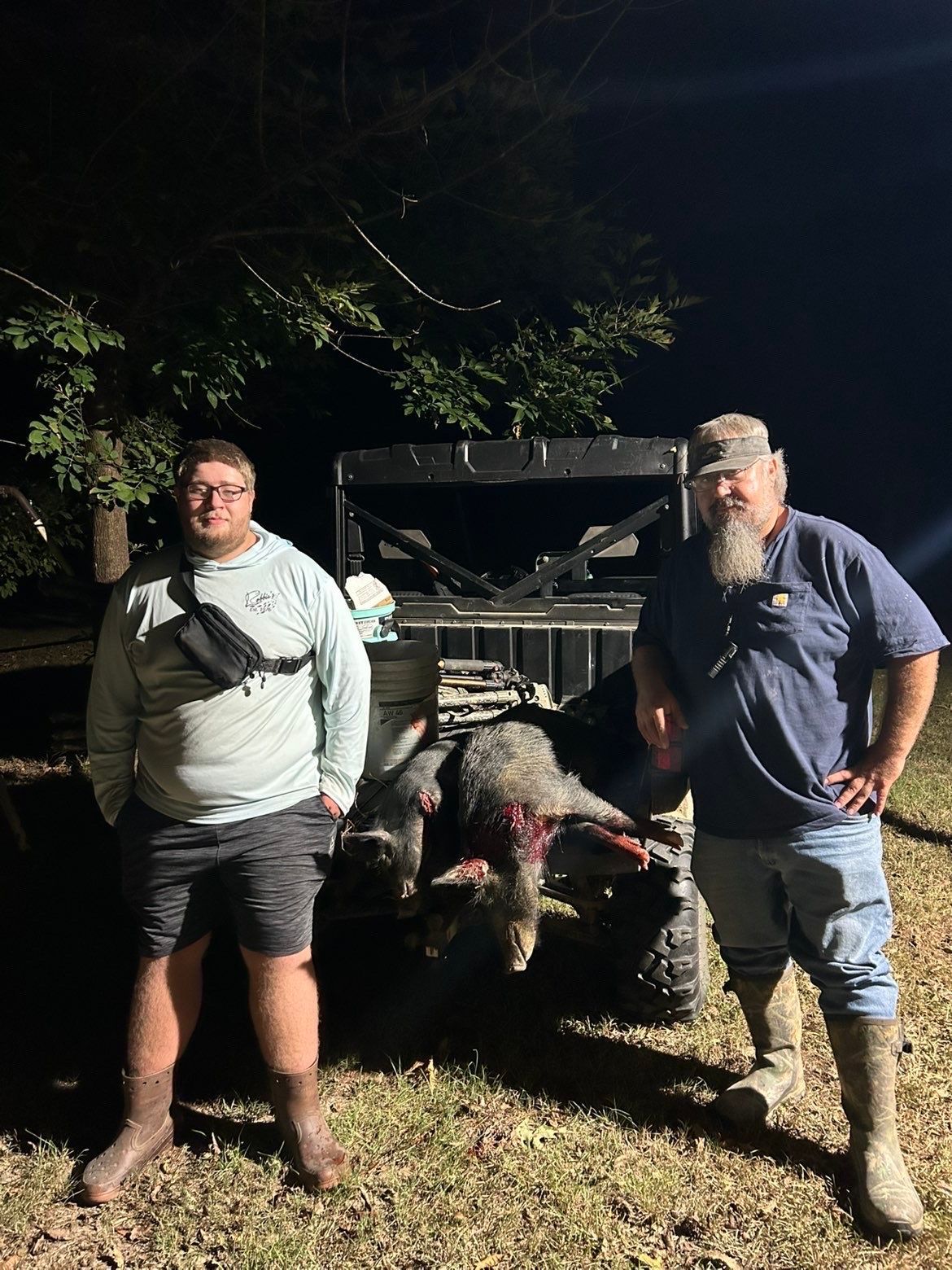 Two people stand near a dead wild boar in front of an off-road vehicle at night.