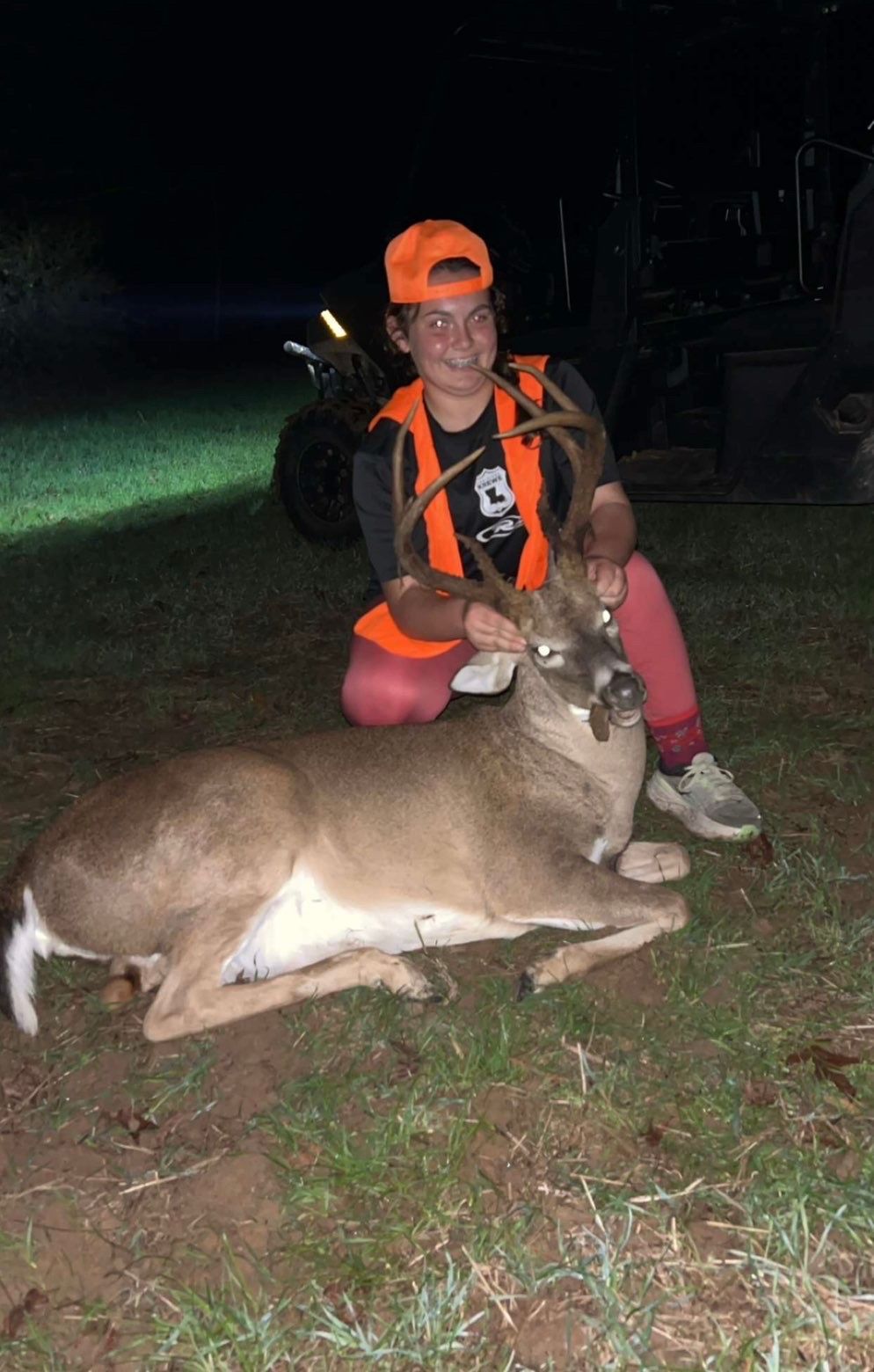 Person in orange hunting vest kneels beside a large deer, posing outdoors at night.