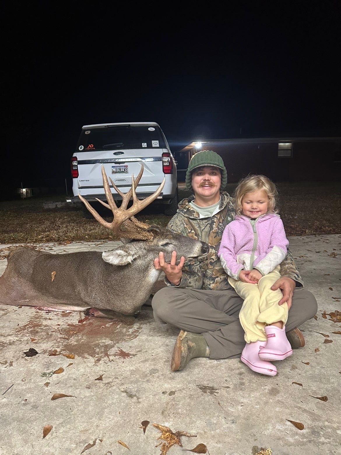 Man and child seated with a large buck. Outdoors at night, truck in background. Man wearing camo, child in pink.
