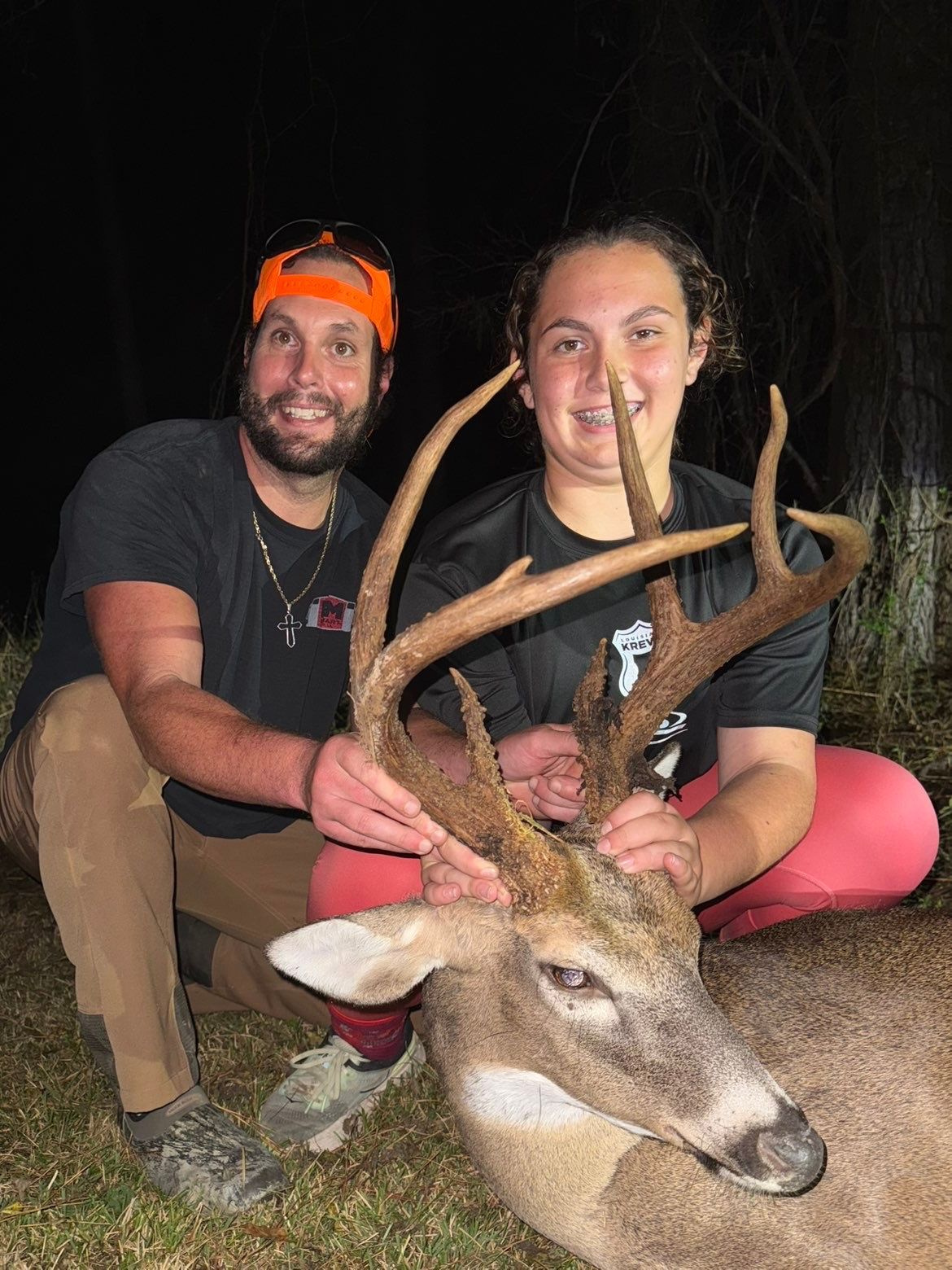 Two people kneeling with a deer, holding its large antlers. Dark setting, orange headband, smiling.