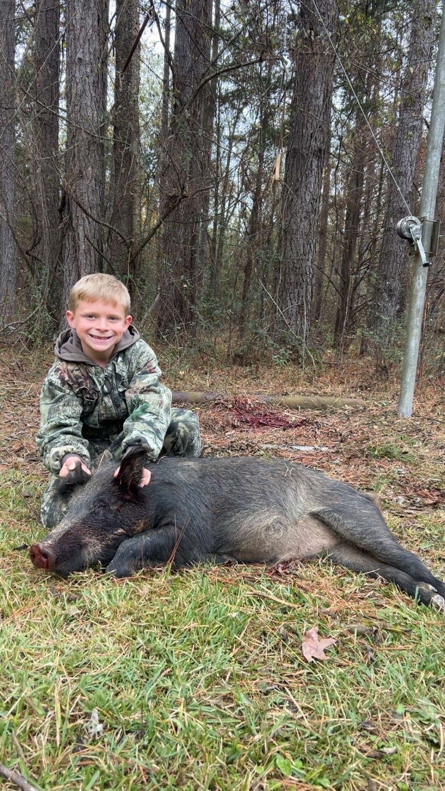 Boy in camouflage kneels beside a wild boar he has hunted. Outdoors, surrounded by trees.