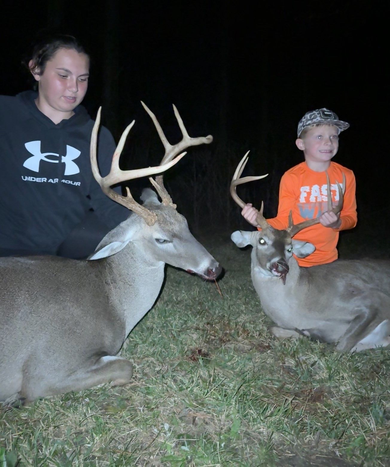 Two deer with antlers lie on the grass, flanked by a person and a child, at night.