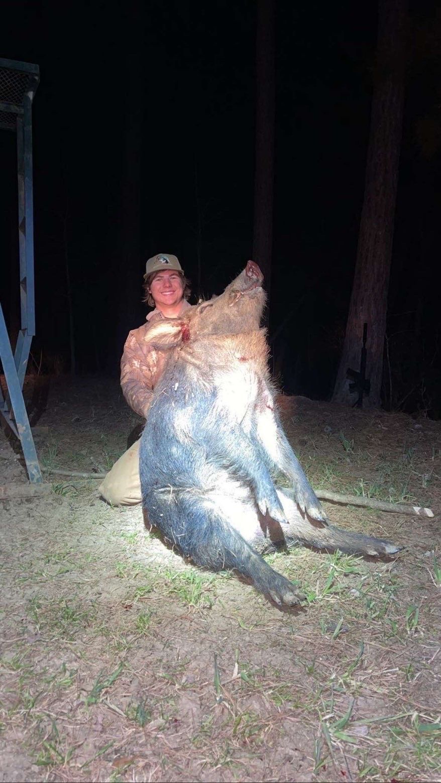 Hunter kneels beside a large, dark-haired wild hog. Both are lit by an artificial light source at night.