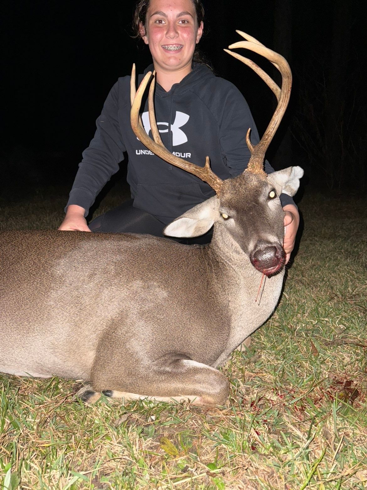 Person kneels beside a large deer with impressive antlers, both on grass at night.