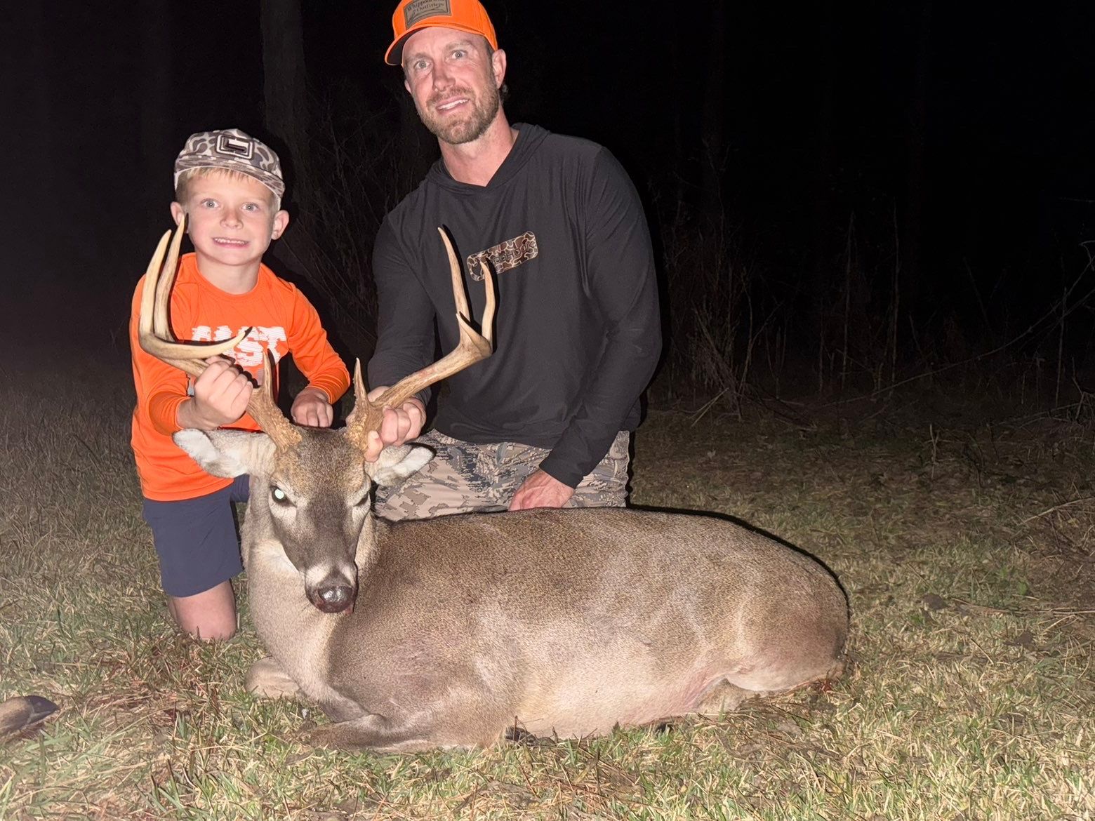 Man and child with a large buck. They are outdoors at night, smiling. The child holds the antlers.