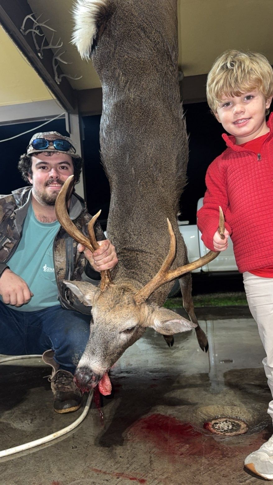 Man and child with a deer carcass. Man holds antlers, child holds a knife, both smiling. Outdoors, concrete.