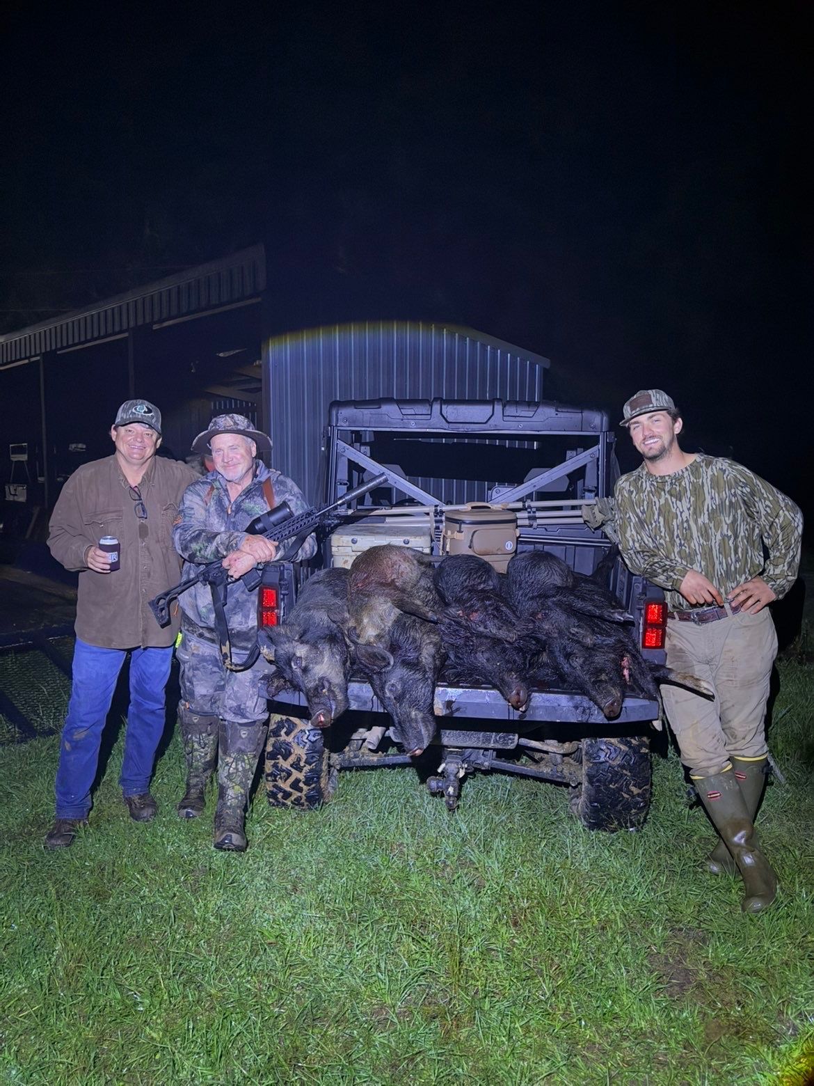 Three people stand beside a truck bed filled with hunted animals at night.