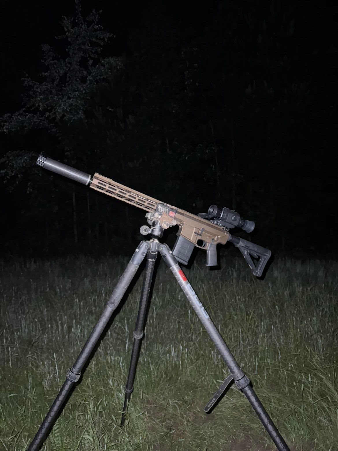 Rifle with scope and suppressor mounted on a tripod in a grassy field at night.