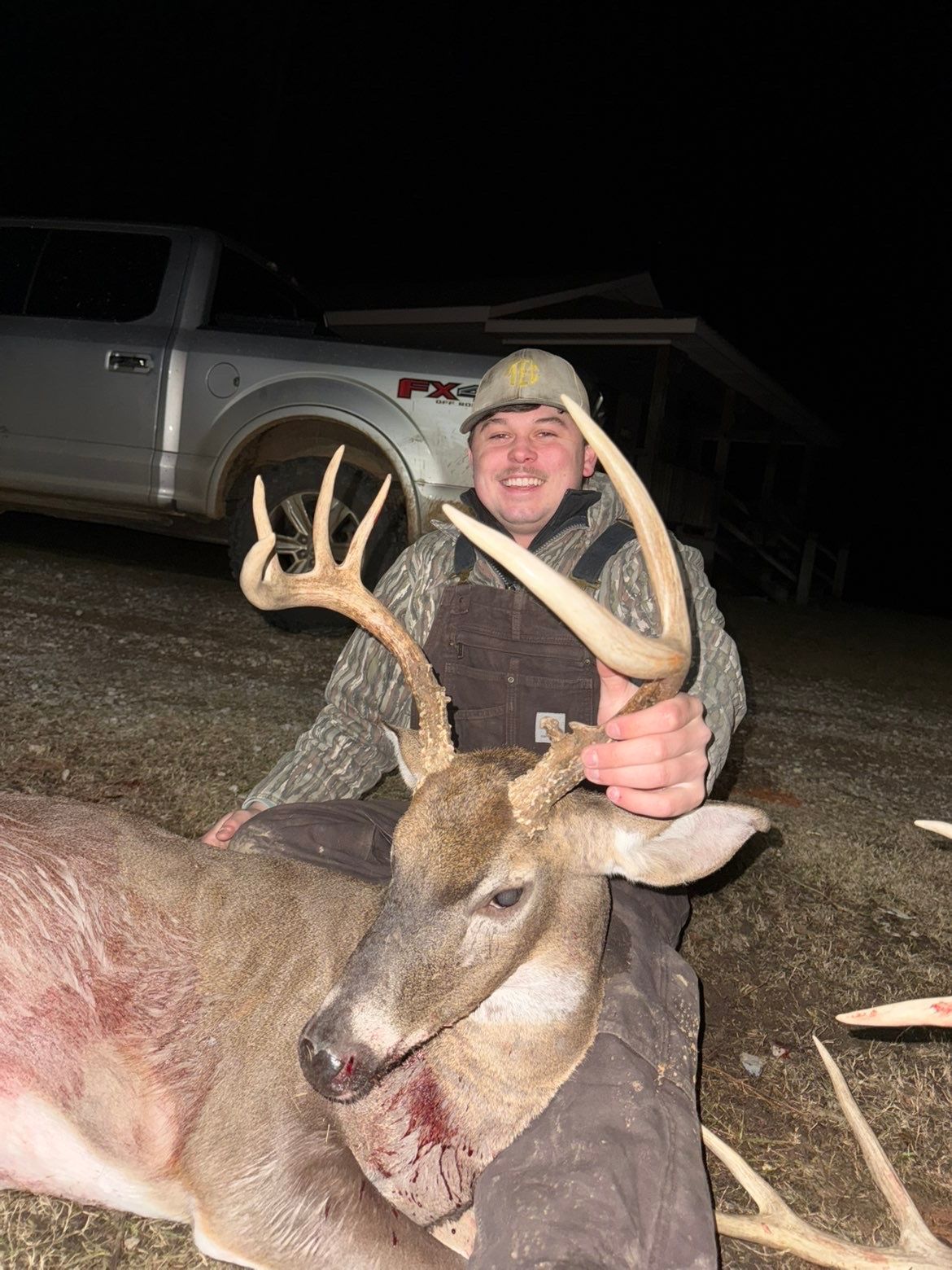 Hunter kneeling with a harvested deer at night, smiling and holding its antler. A truck and building are in the background.