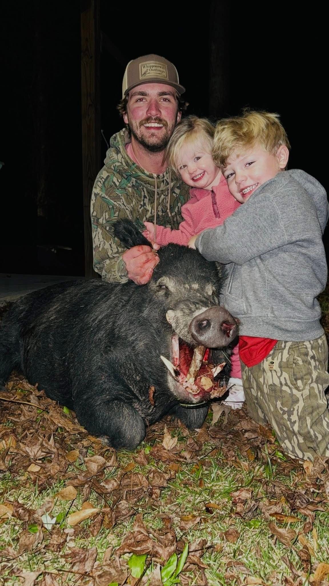 Man and two children with a dead wild boar. Man smiles, children hug boar. Outdoors, at night.