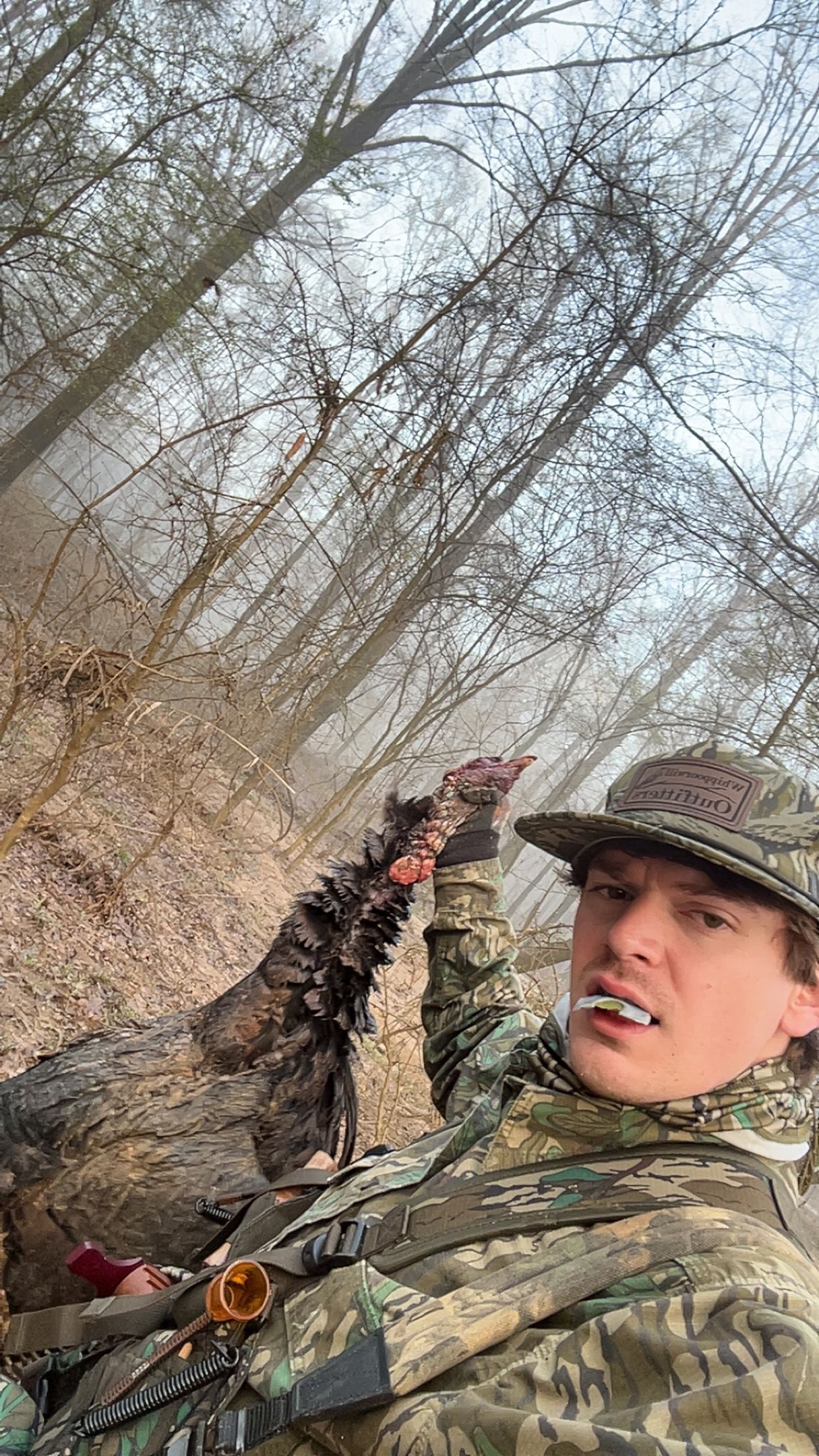 Man in camouflage holds a dead turkey's leg in a wooded area. He wears a hat and has a cigarette in his mouth.