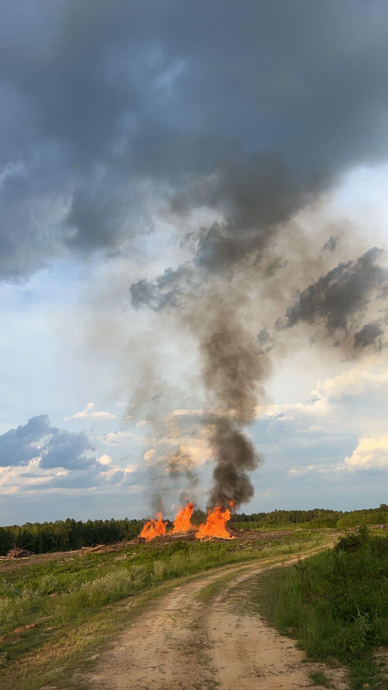 Flames and black smoke billow from a fire on grassy land with a dirt path, under a cloudy sky.