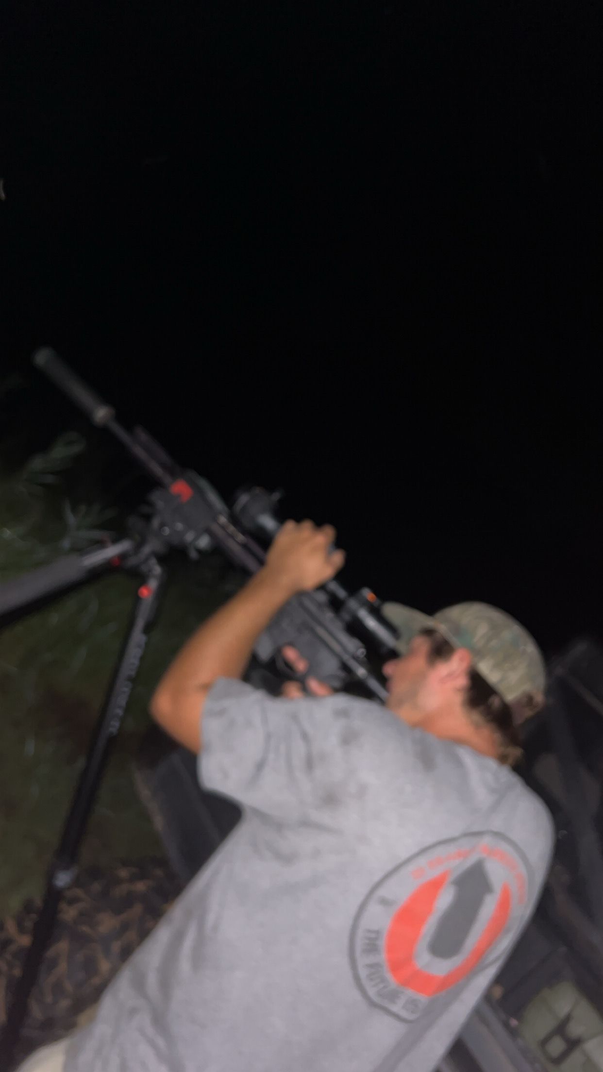 Person aiming rifle with scope on tripod at night.