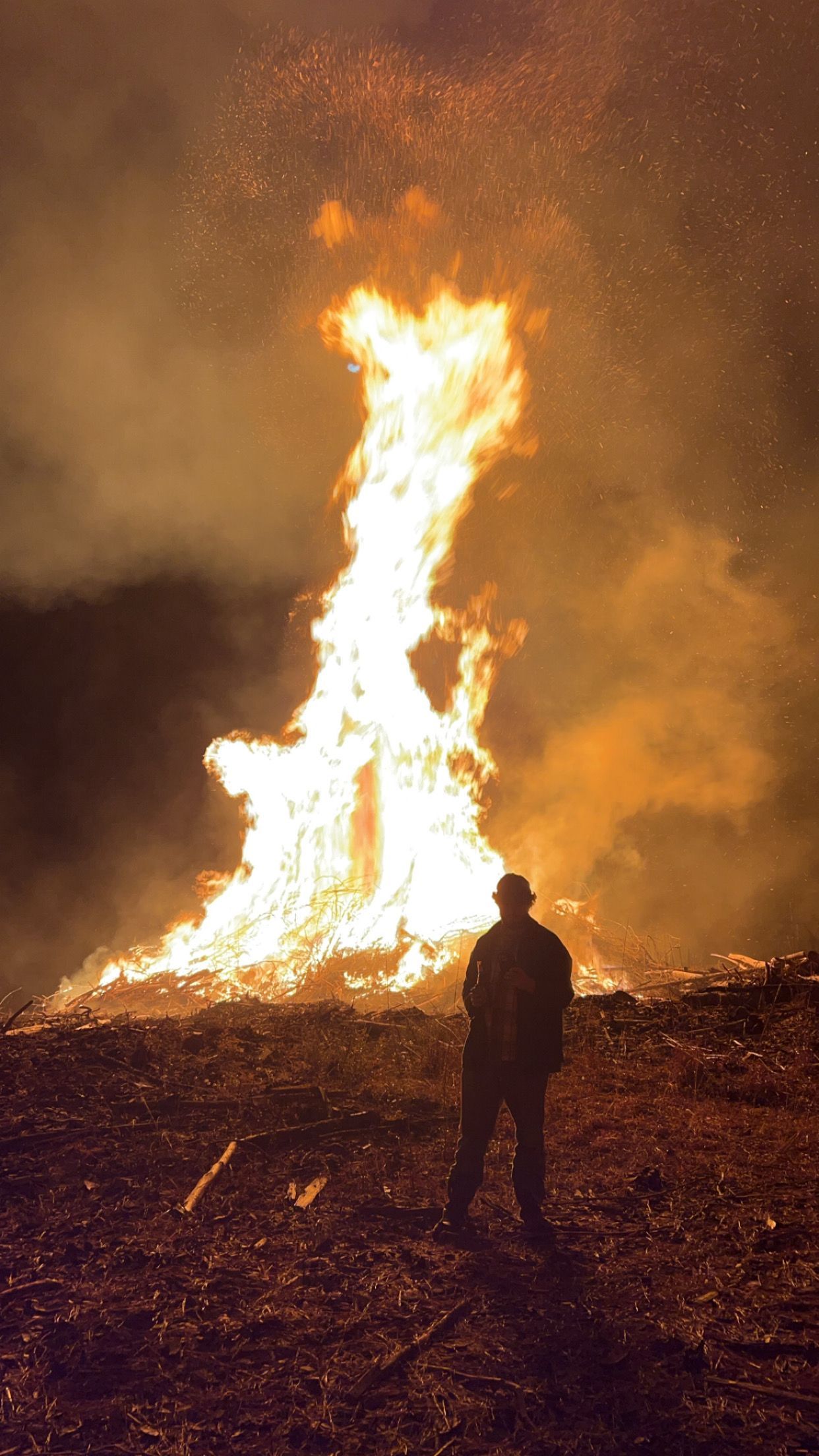 Person watching a large bonfire at night, flames reaching high, sparks in the air, dark surroundings.