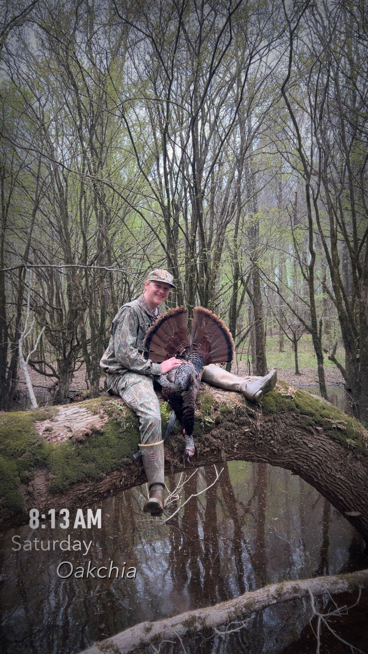 Hunter sitting on log over water, holding a turkey in the woods.
