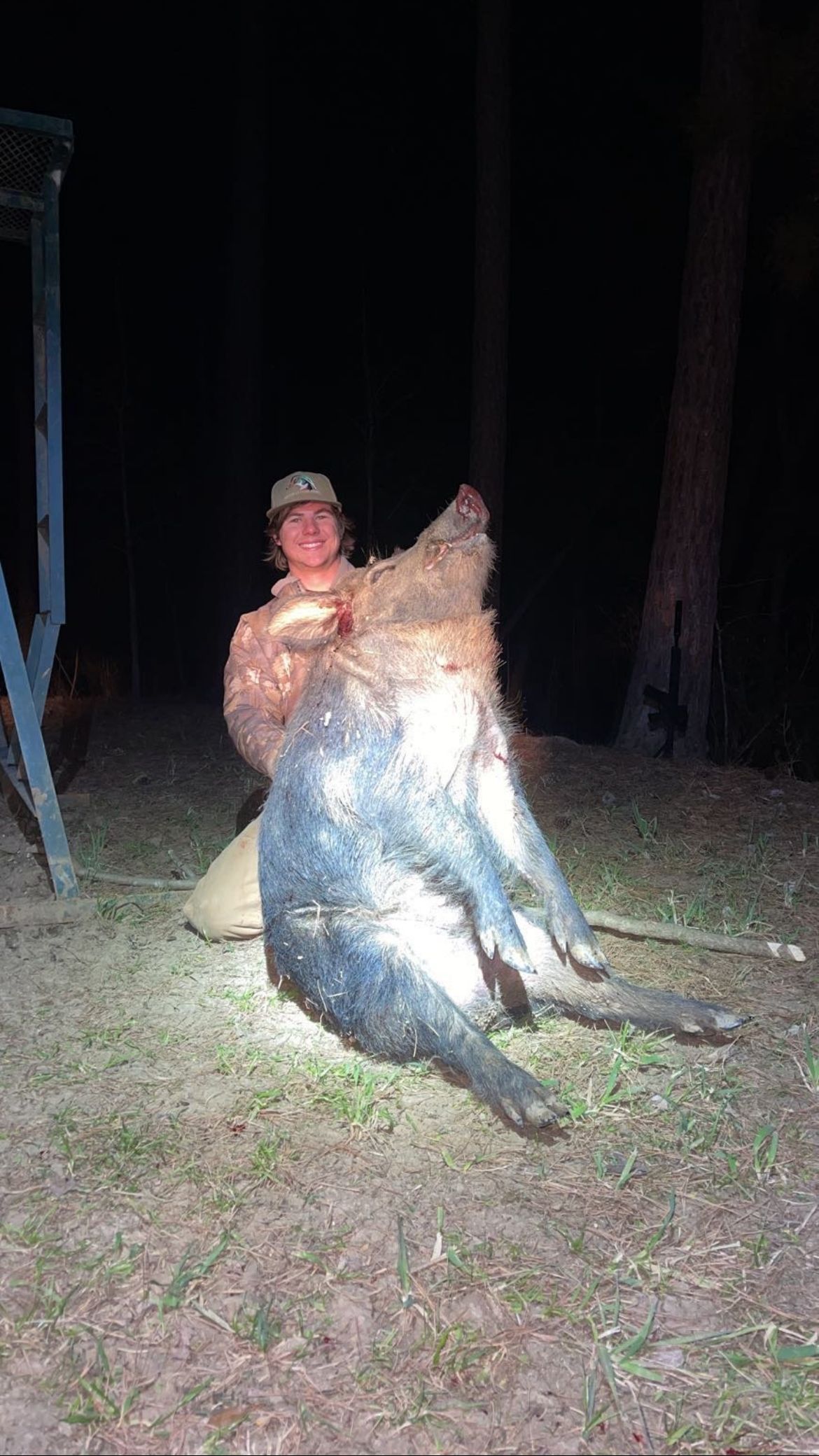 Person kneeling, holding a large, dark wild boar carcass in a dark, outdoor setting.