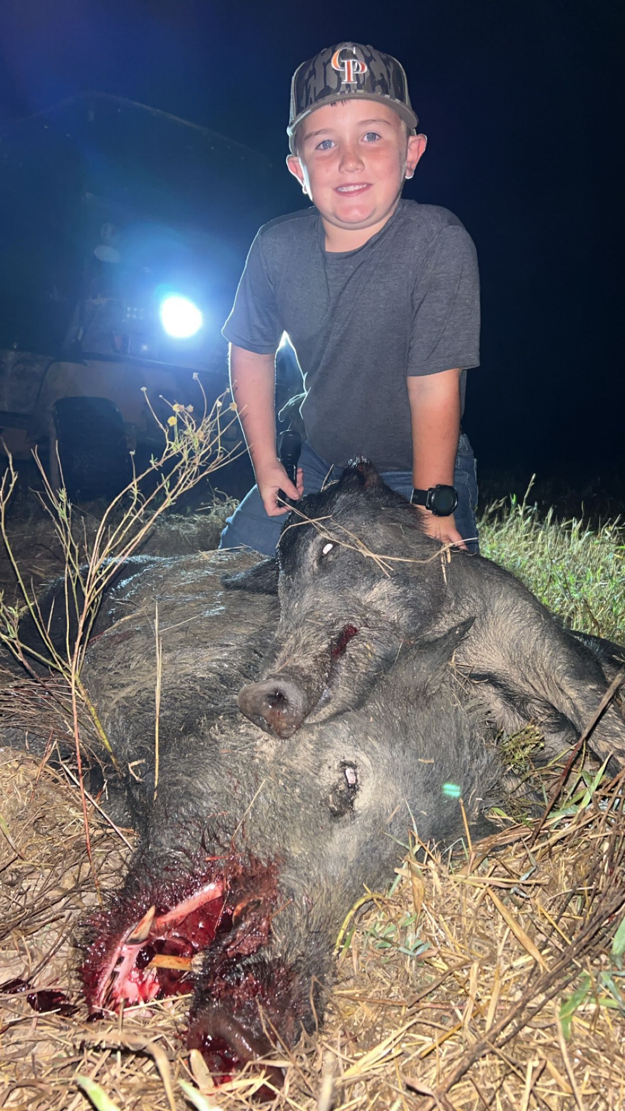 Boy on a large, dead pig at night, blood visible. He wears a hat and smiling.