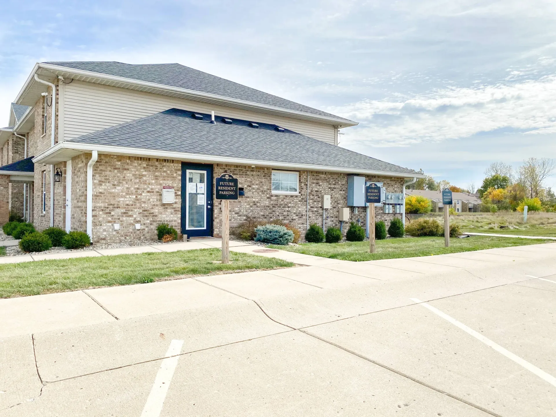 Exterior of a brick multifamily leasing office with sidewalk, shrubs, and future resident parking signs.