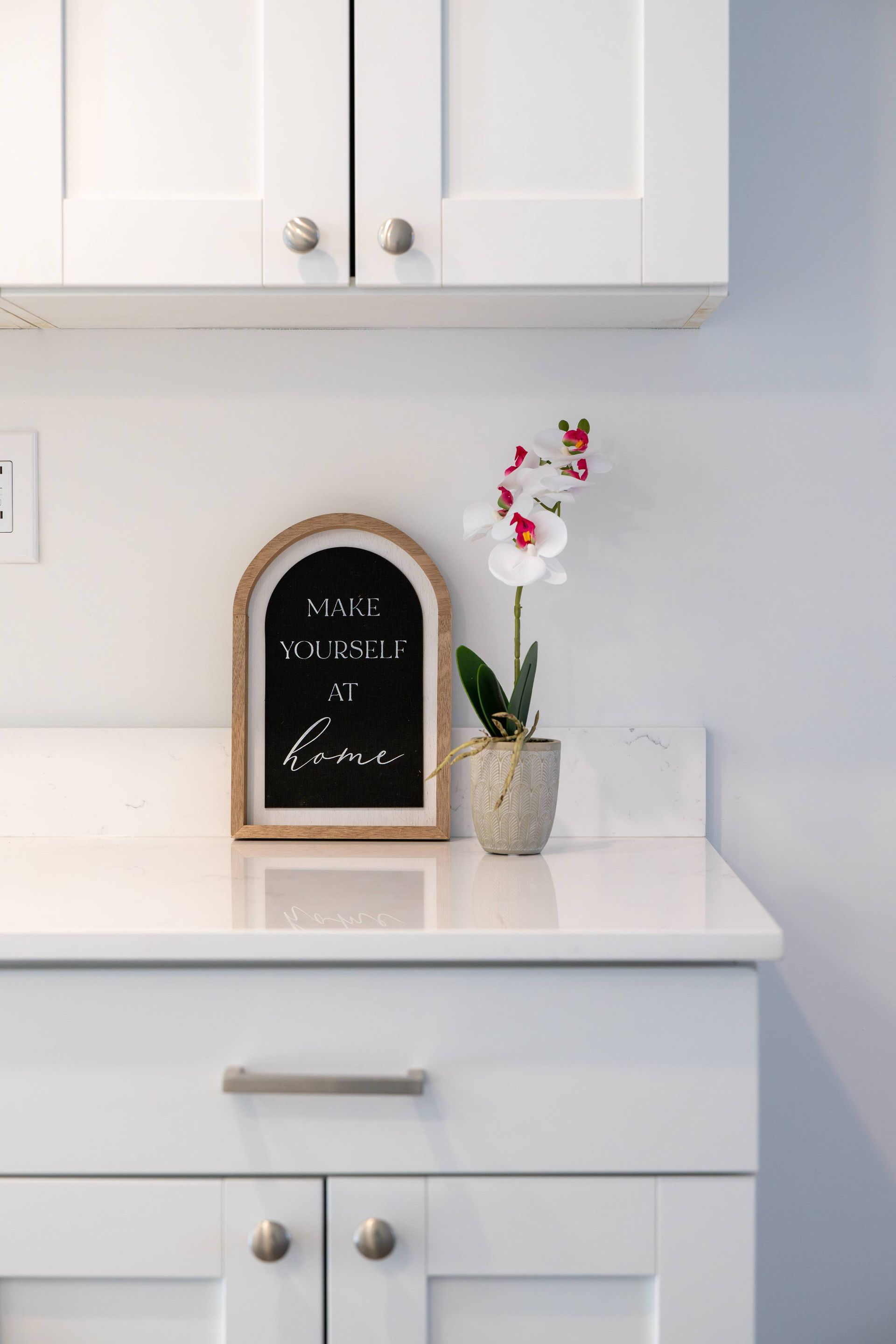 White kitchen counter with cabinets. Orchid and framed sign that reads