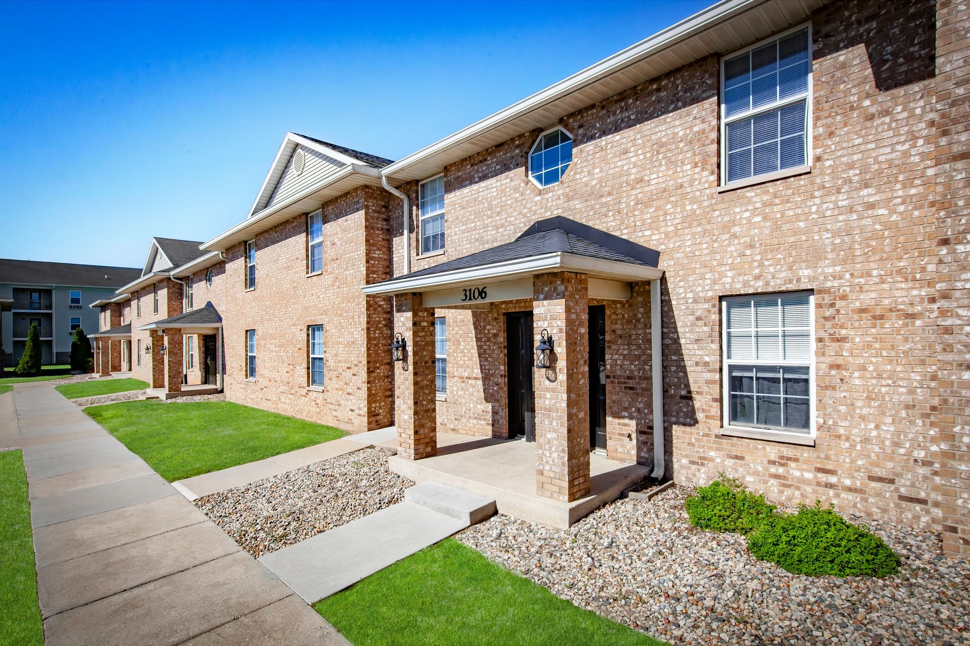 Brick apartment building entrance with covered walkway, white trim, and landscaped lawn