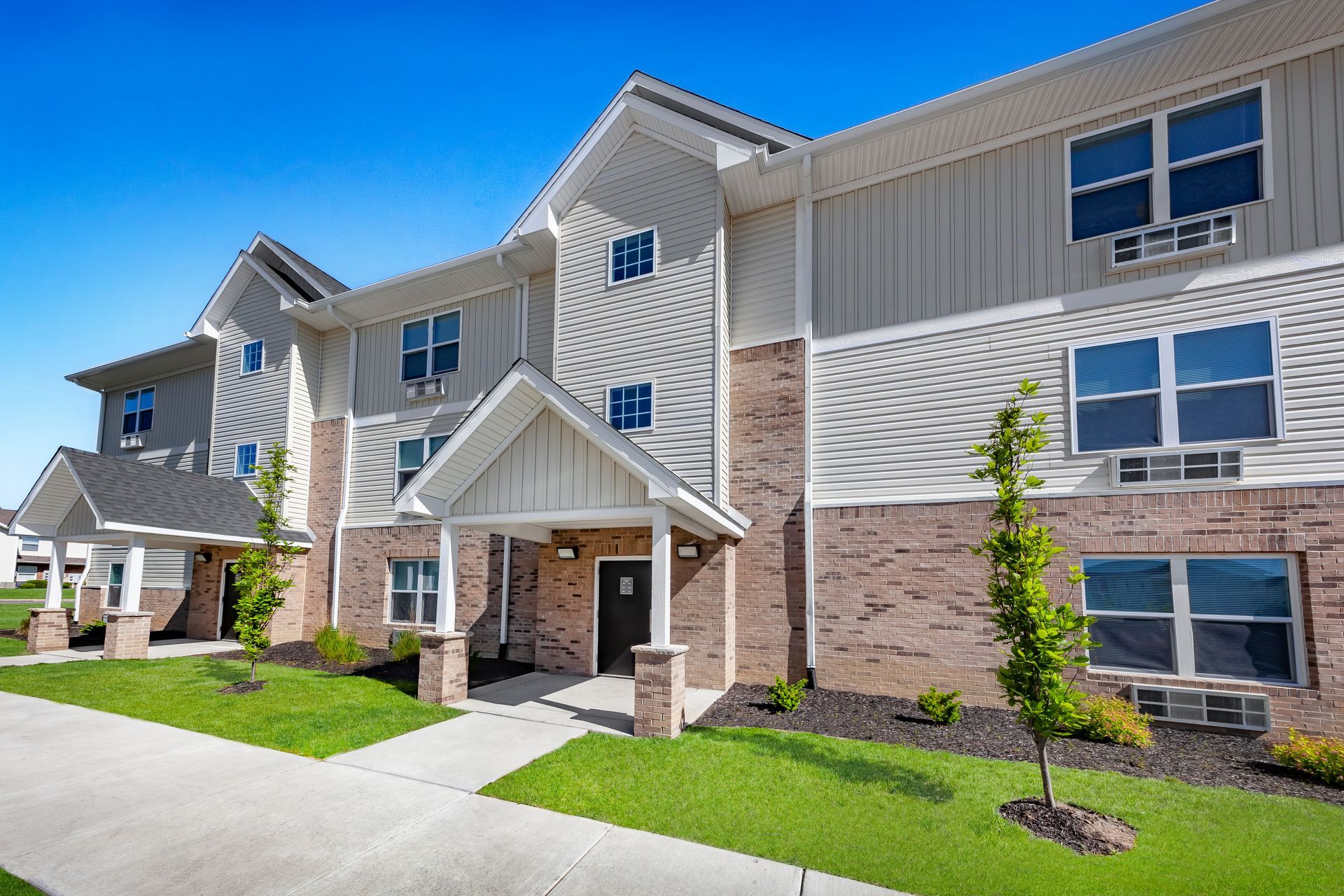 Modern apartment building exterior with beige siding, stone accents, large windows, and landscaped lawn